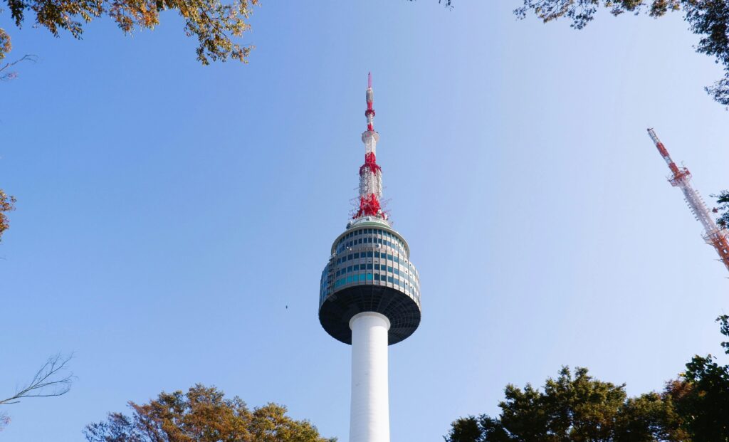pexels-photo-34416792-34416792 Namsan Seoul Tower rises into a clear blue sky surrounded by autumn foliage.